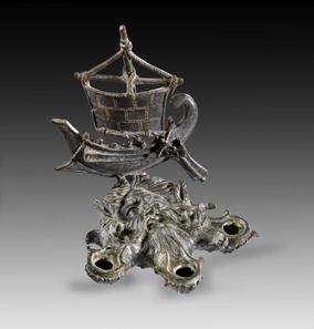 Nr. 234: Bronzene Lampe in Gestalt des Oceanus mit Schiffsaufsatz. Römisch, 1.-2. Jh. n. Chr. Lampe in Form eines bärtigen Oceanus-Kopfes mit Krabbenscheren. Am Kopfende Aufsatz in Form einer römischen Galeere. Aus Sammlung Shlomo Moussaieff, seit 1948, Israel. Mit Ausfuhrgenehmigung der israelischen Antikenbehörde. Schwarzgrüne Patina, Mast und Segelecke gebrochen, Mast mit Stift repariert, Mastspitze fehlt, Teile der Taue fehlen, kleines Loch auf einer Schnauze. Schätzung: 50.000,- Euro. Zuschlag: 55.000,- Euro.